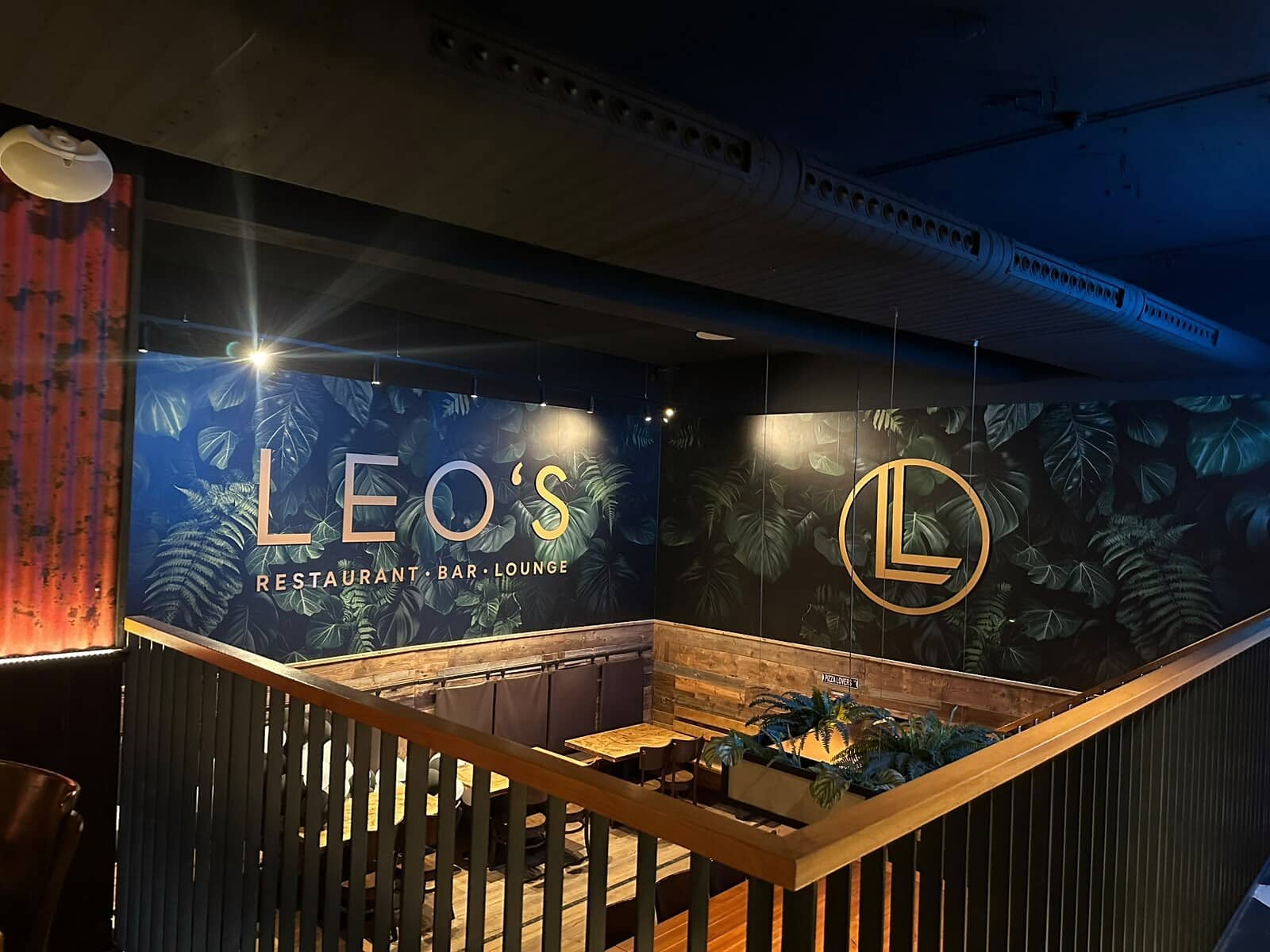Лаунж в Leo's: стена из зелёной листвы с золотой надписью и монограммой Leo's, перед ней деревянные скамьи и растения.