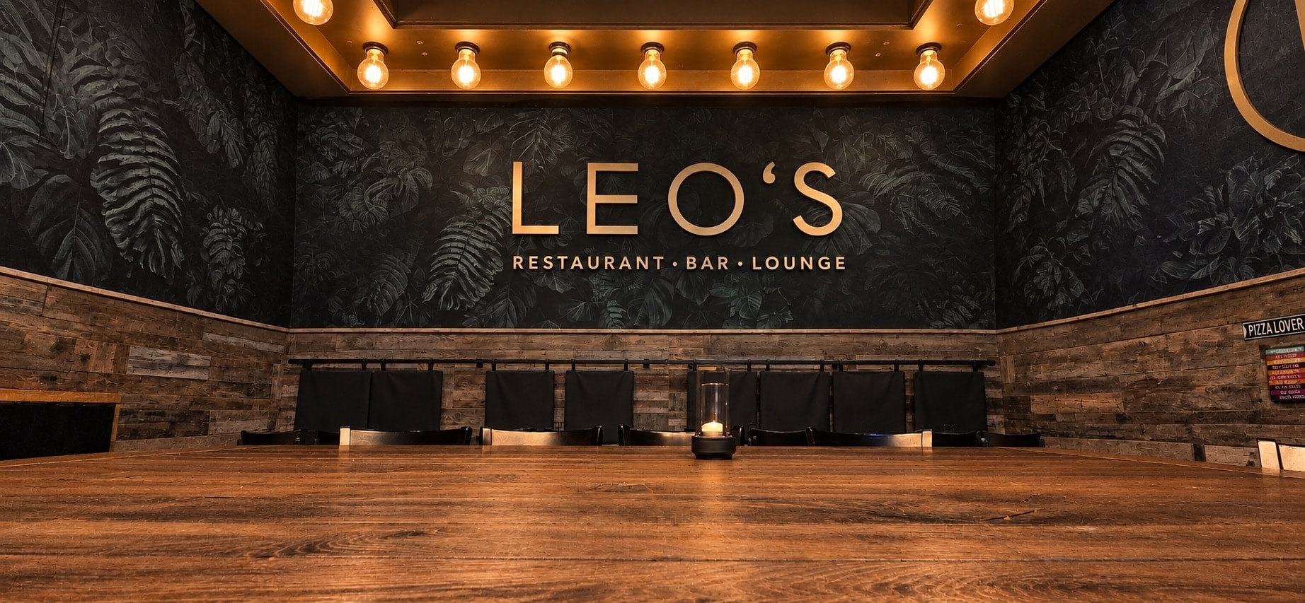 Der Gastraum im Leo's: eine dunkle Blätterwand mit dem goldenen Schriftzug „LEO'S – Restaurant · Bar · Lounge“, darüber eine Reihe Glühbirnen, vorne ein langer Holztisch mit Windlicht.